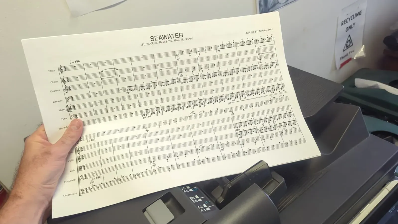 A hand holding a freshly printed page of the SEAWATER score beside a desktop printer.
