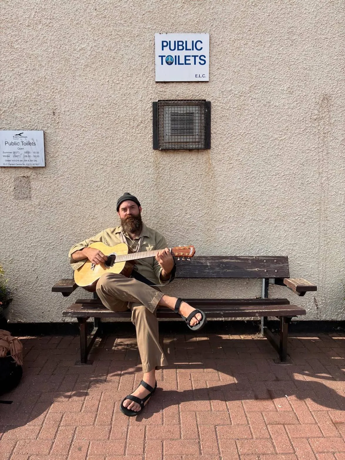 Nicholas Daly on a wooden bench playing an acoustic guitar, in a knit cap and sandals, under a Public Toilets sign on a rendered wall.